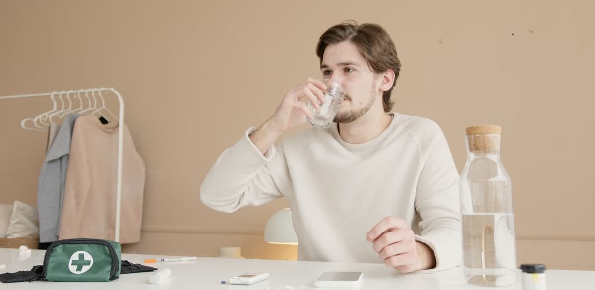 Man in white long sleeves drinking water from glass at home with medical supplies.
