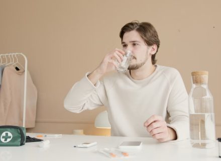 Man in white long sleeves drinking water from glass at home with medical supplies.