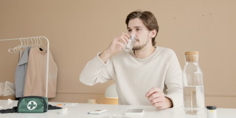 Man in white long sleeves drinking water from glass at home with medical supplies.